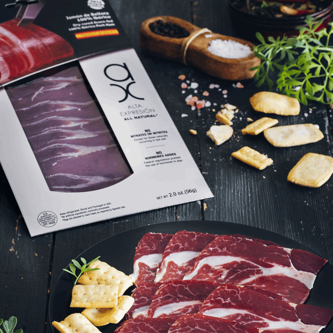 a plate with slices of Jamon Iberico, a grey package and some crackers on a black table